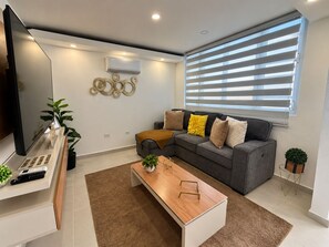 Uma sala de estar moderna com sofá em forma de L, mesa de centro de madeira, TV de tela plana e uma janela grande com persianas.