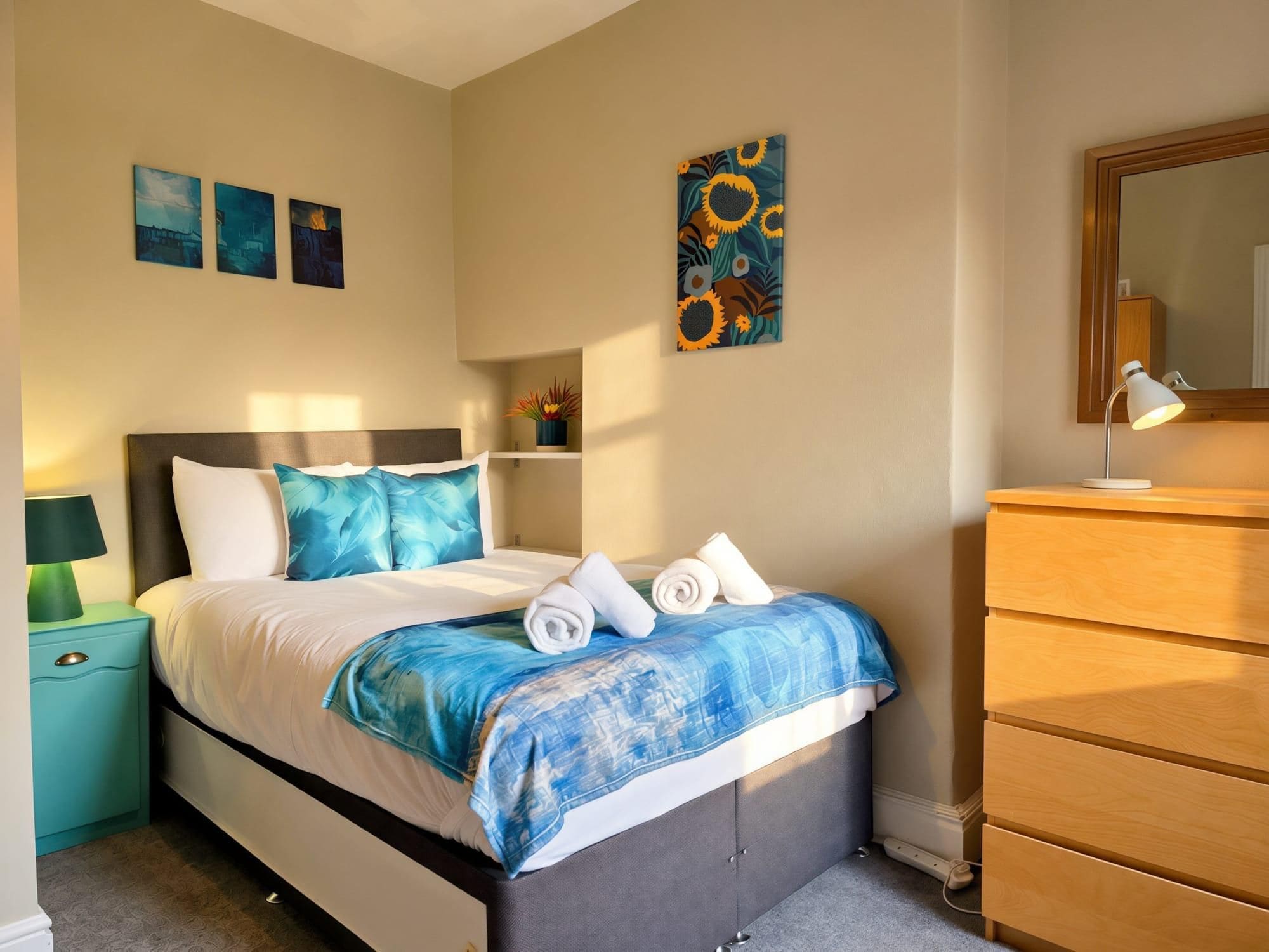 A neatly arranged bedroom with a bed, bedside table, dresser, and artwork on the walls.
