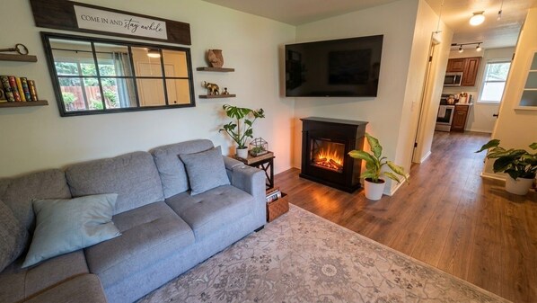 A living room with a grey sofa, a fireplace, a flat-screen TV, and a kitchen in the background.