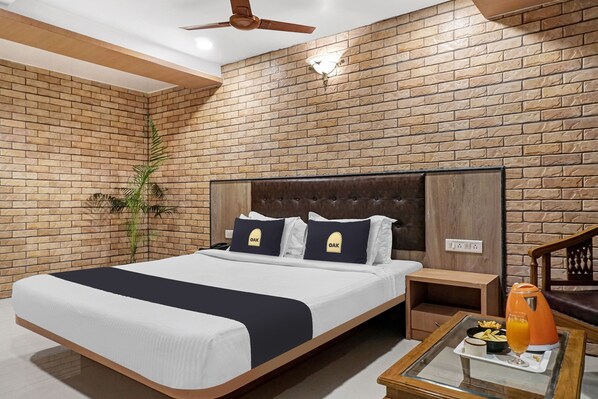 A hotel room with a brick-patterned wall, a bed with a dark headboard, two pillows with 'OAK' text, a nightstand, a glass table with a tray of food and drink, and a ceiling fan.