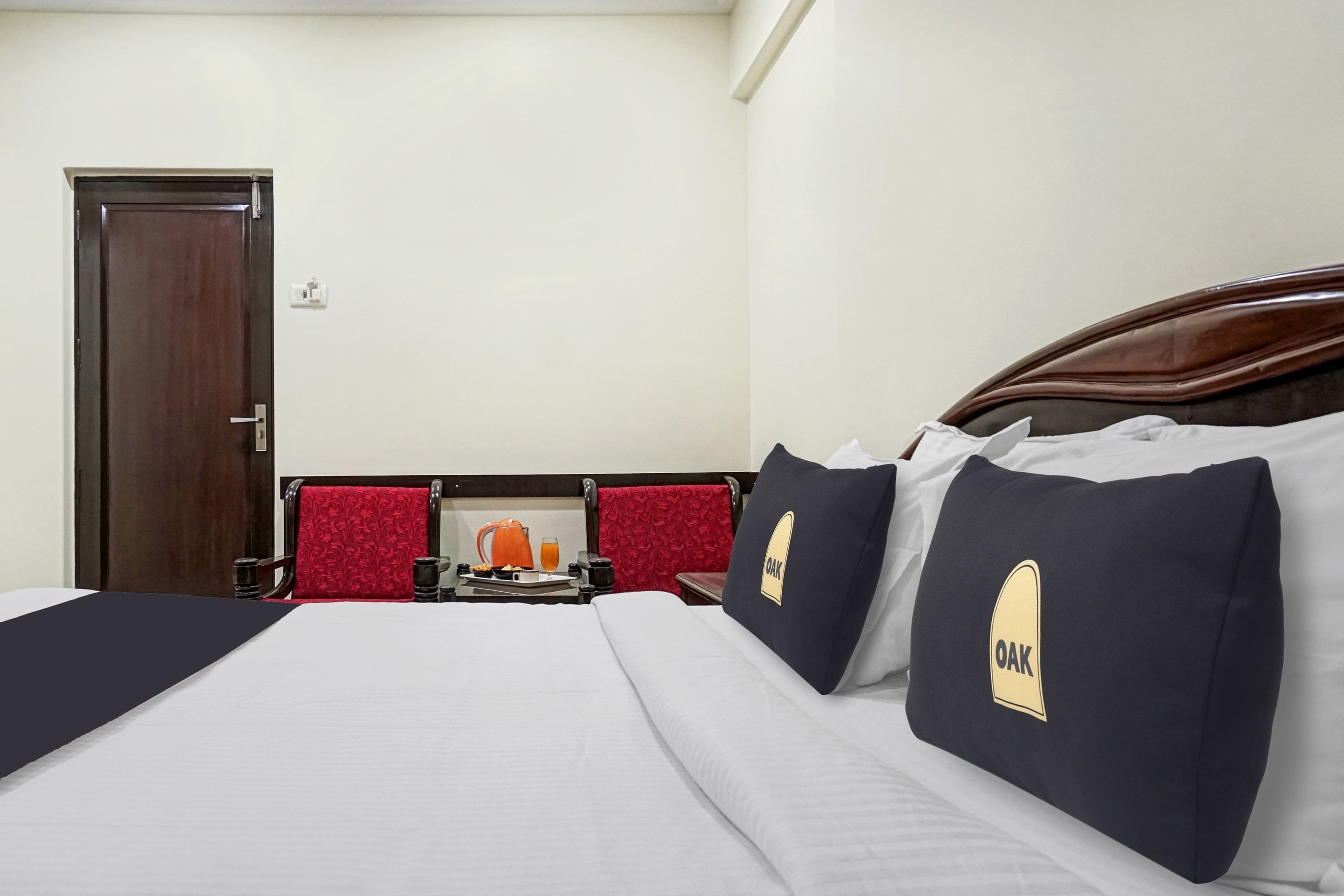Habitación de hotel con una cama, una puerta de madera, una silla roja y una pequeña mesa con una tetera y un vaso de jugo de naranja.