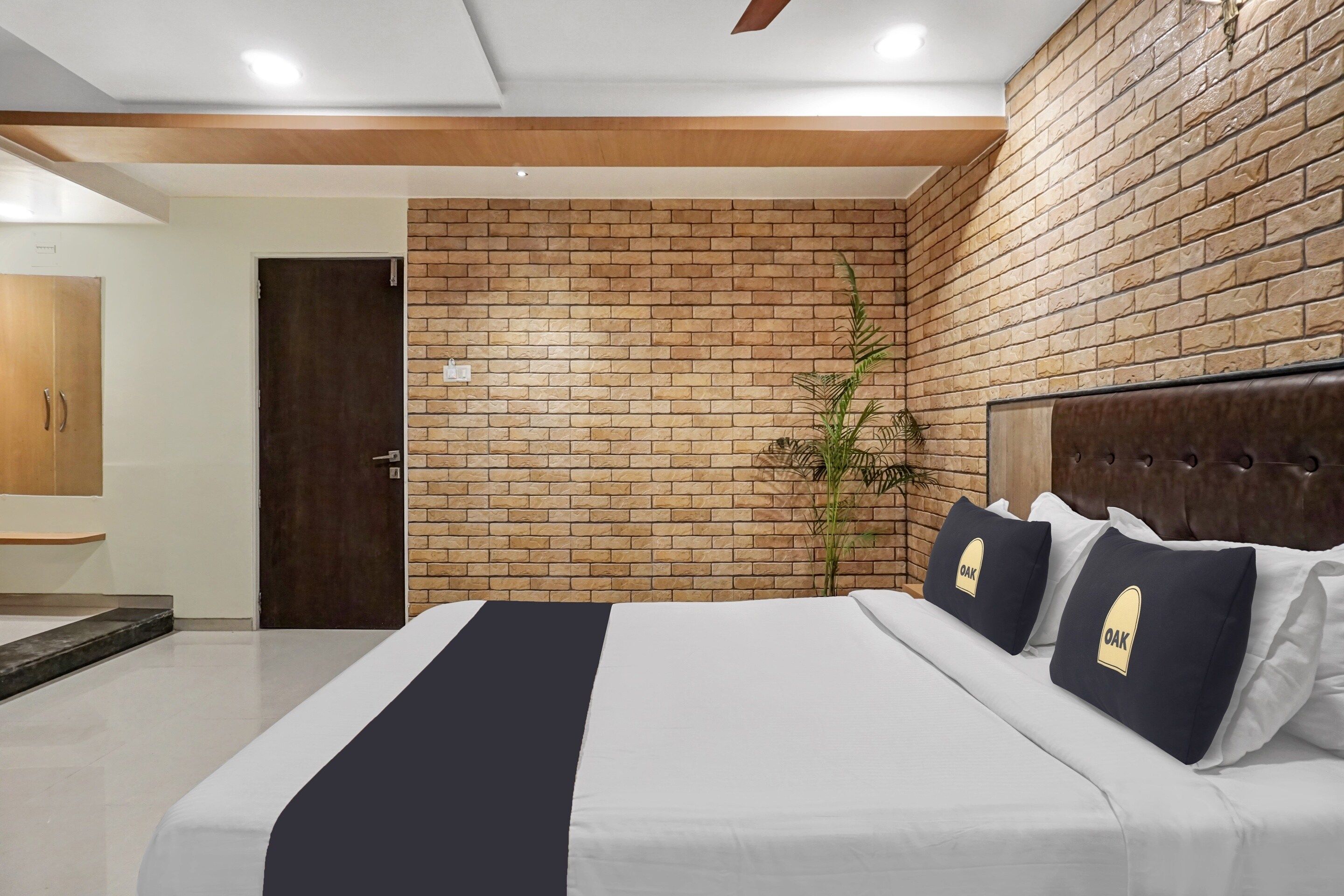A hotel room with a brick wall, a bed with white bedding and dark pillows, a wooden door, and a plant.