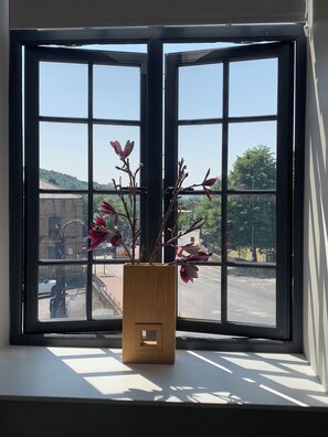 Una ventana con un florero de madera que contiene ramas y hojas, con vista a una calle y edificios.