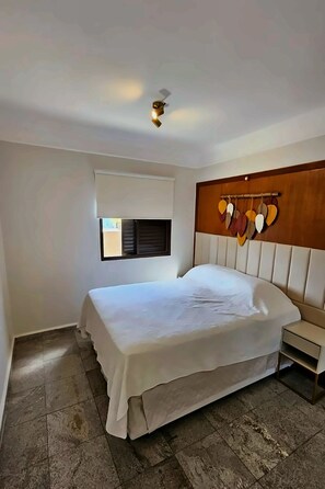 Quarto com cama, mesa de cabeceira, luminária na parede e janela com persianas.