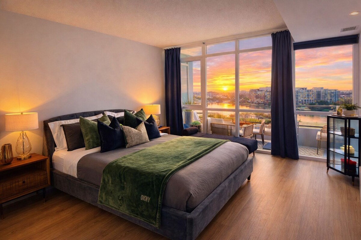A modern bedroom with a large bed, a nightstand, a lamp, and a view of the city at sunset.