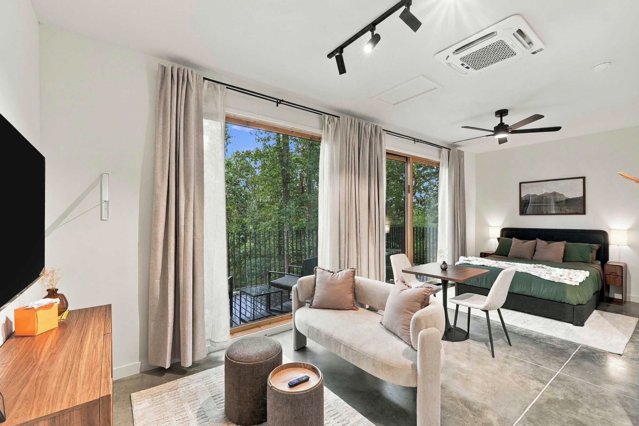 A modern living room with a sofa, a dining table, and a balcony with a view of trees.