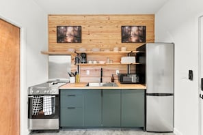 A compact kitchen with a built-in oven, a microwave, a stainless steel refrigerator, and wooden shelves above the countertop. - The Hummingbird | Bentonville Woods + Trails (Bentonville)