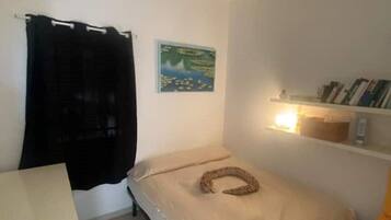 Quarto pequeno com uma cama de solteiro, roupa de cama branca, cortina preta, um quadro na parede e uma estante com livros.