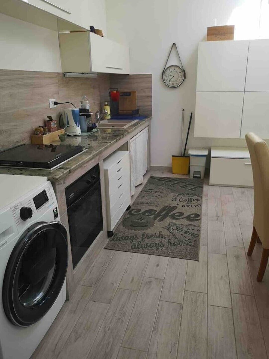 Een compacte keuken met een wasmachine, een koffieapparaat en een klok aan de muur.