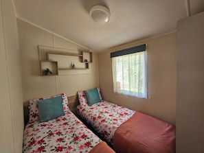 Quarto com duas camas, cada uma com roupa de cama floral e uma única almofada. Há uma estante sobre as camas e uma janela com cortinas.