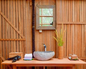 A wooden wall with a window, a sink with a stone basin, a mirror, a potted plant, and a small wooden box.