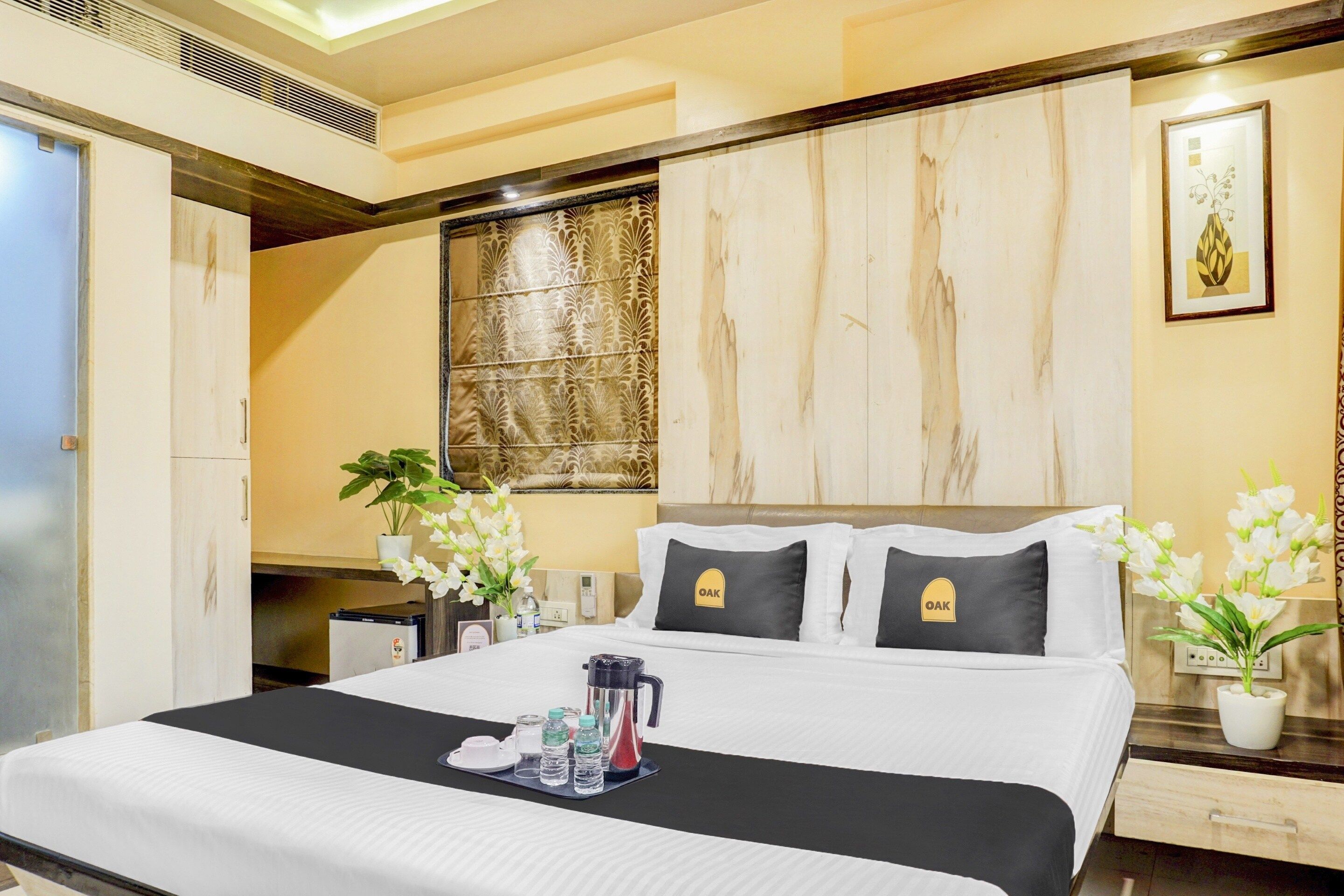 A hotel room with a bed, bedside tables, a nightstand, a wall-mounted heater, a window with a decorative panel, and a framed picture on the wall.