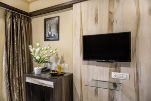 Habitación de hotel con pared de paneles de madera, un televisor de pantalla plana montado en un estante de vidrio, un escritorio con teléfono, un florero con flores blancas y una cortina con estampado de hojas.