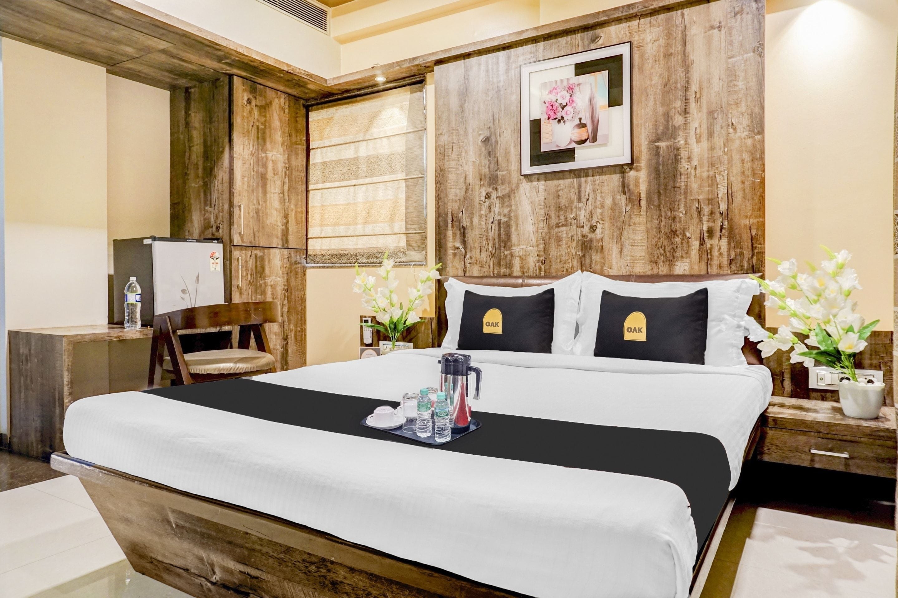 Une chambre d’hôtel avec une tête de lit en bois, un lit avec une literie blanche, une table de chevet avec une lampe, une chaise et un tableau au mur.