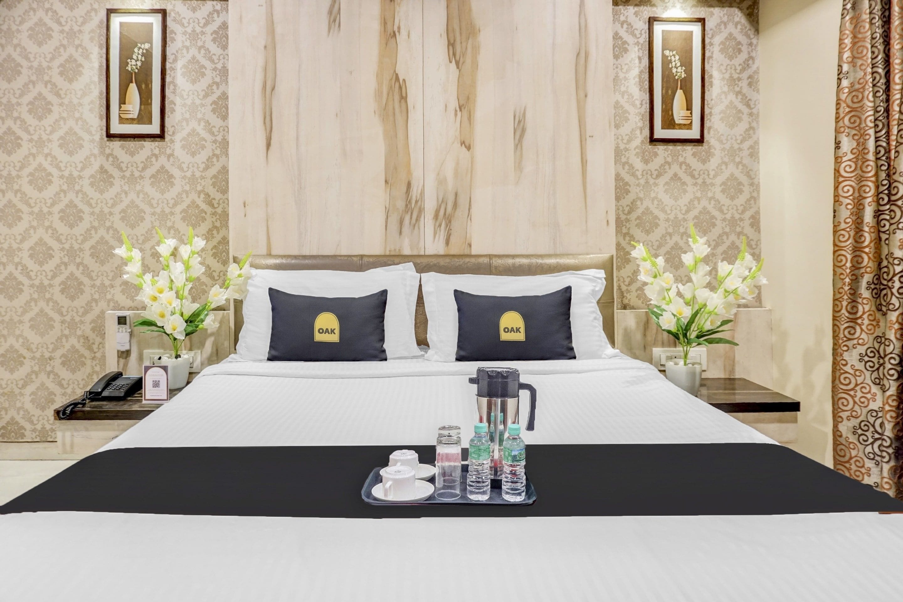 Habitación de hotel con una cama grande, dos almohadas con un logotipo, mesitas de noche con lámparas, un teléfono, dispensador de agua y una bandeja con vasos y botellas.