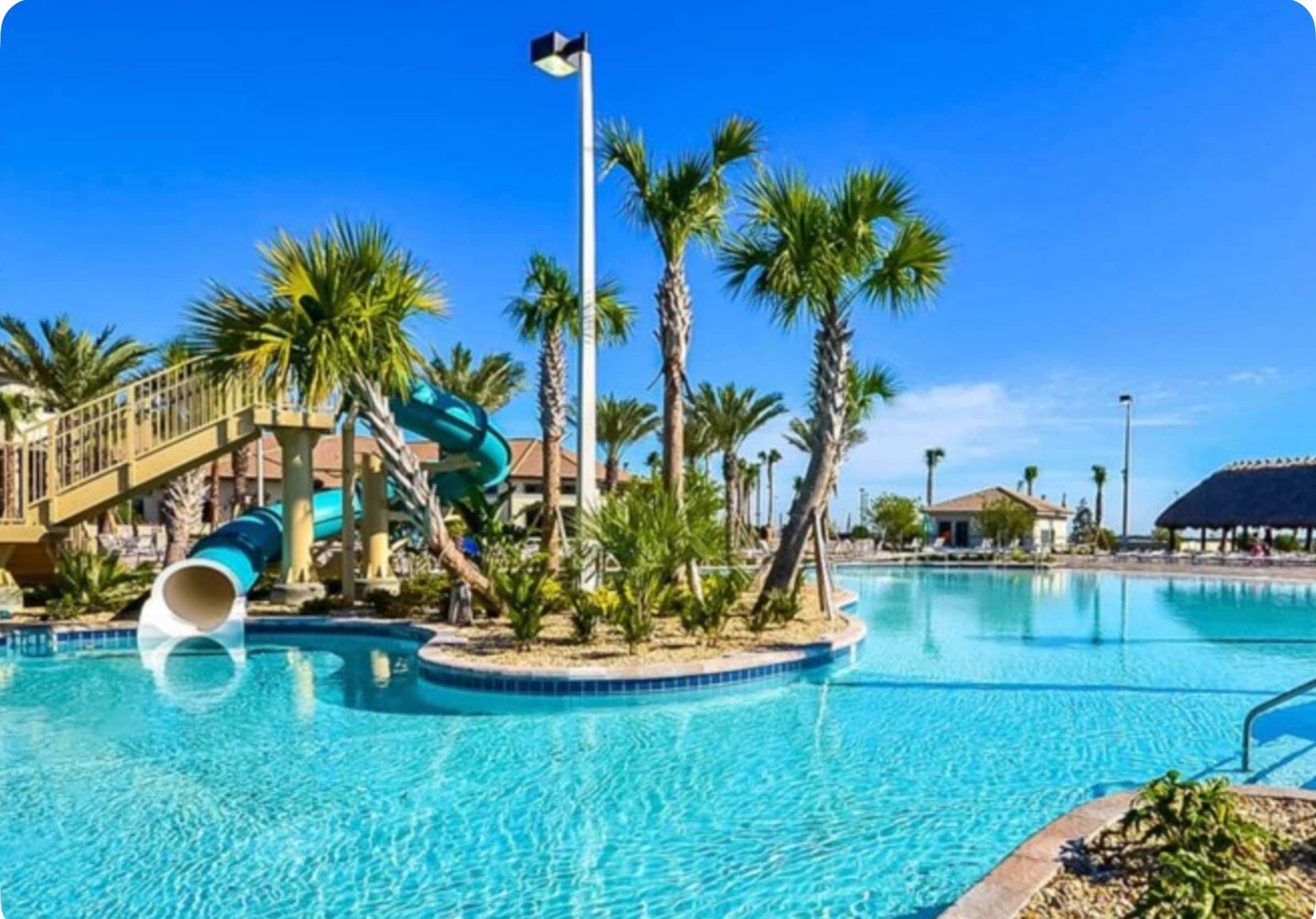 Una piscina con tobogán de agua, palmeras y un cielo azul despejado.