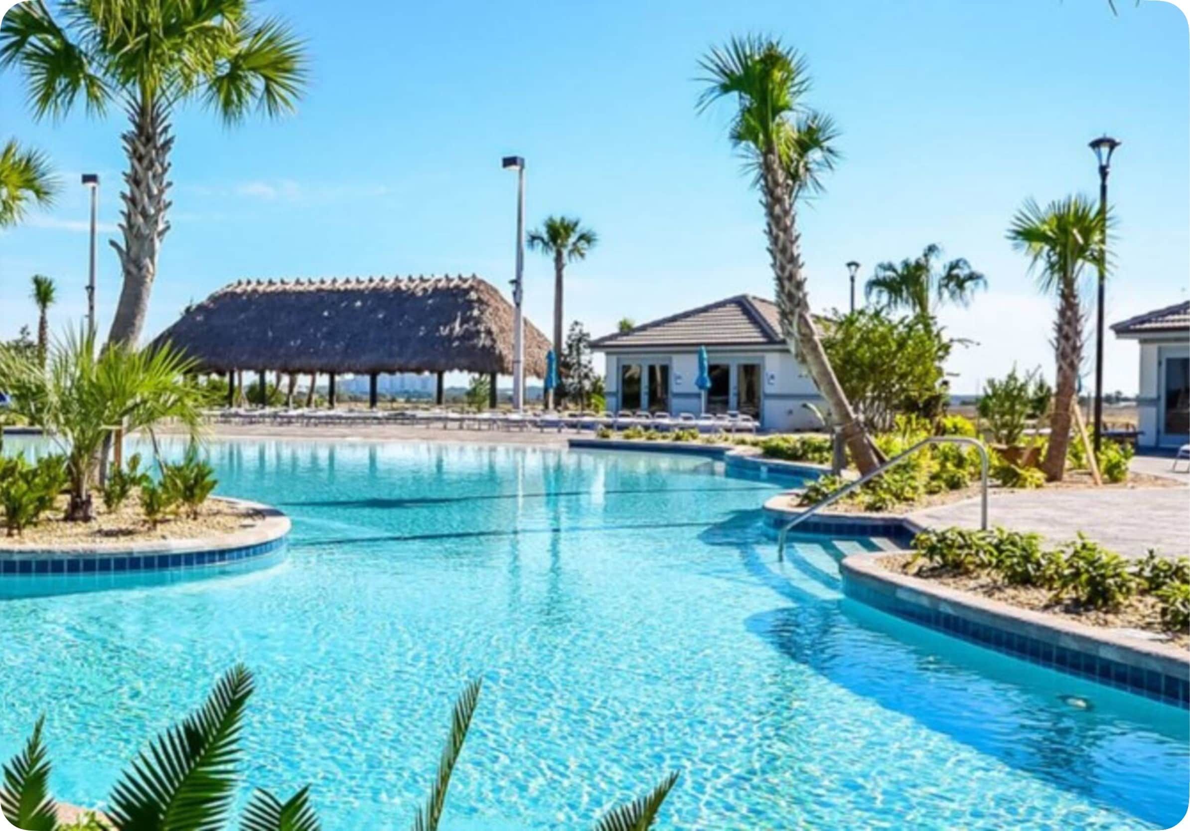 A swimming pool with a thatched-roof structure in the background, surrounded by palm trees and a small building.