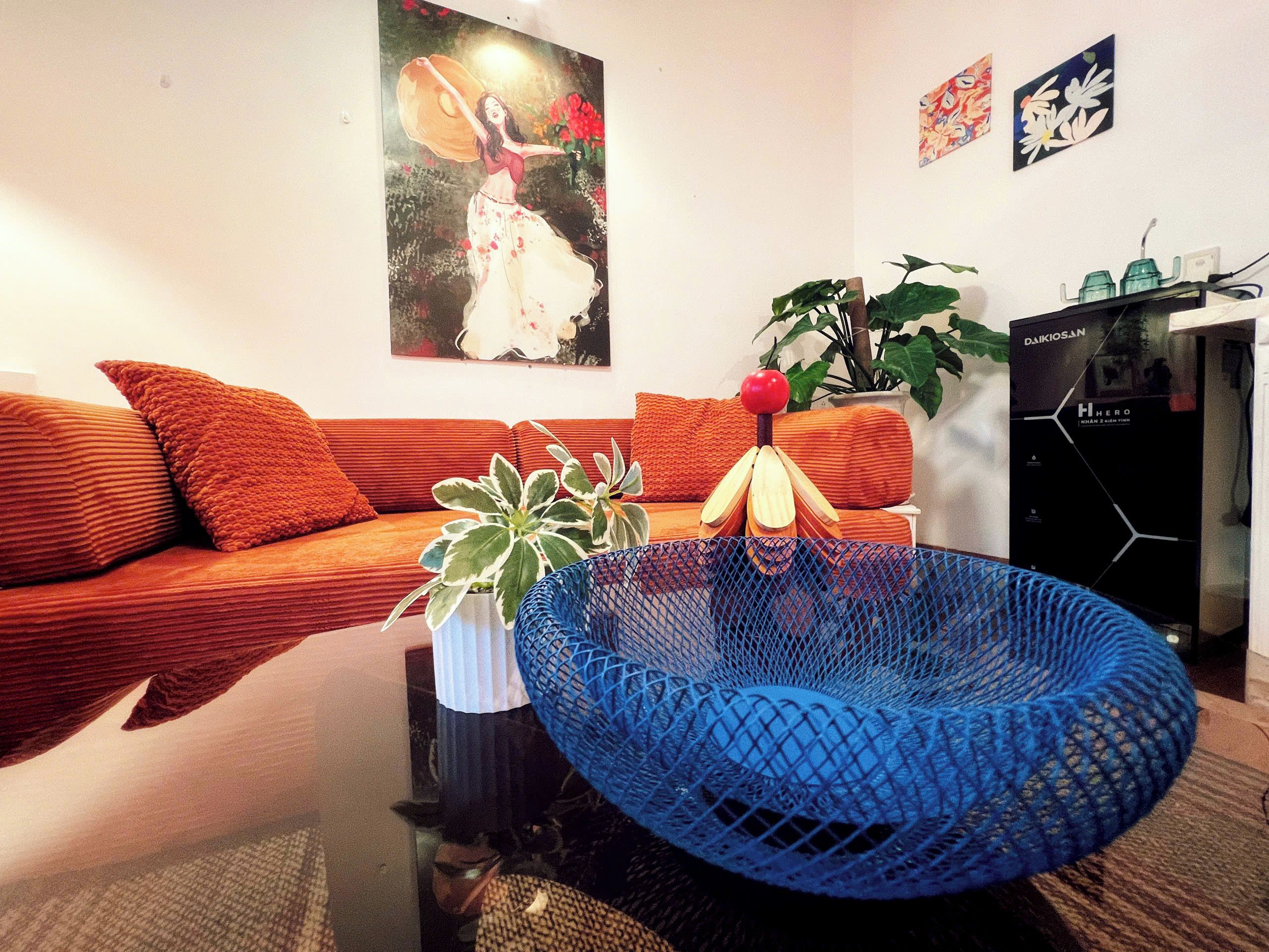 Een woonkamer met een bank, een blauwe geweven stoel, een potplant en een schilderij aan de muur.