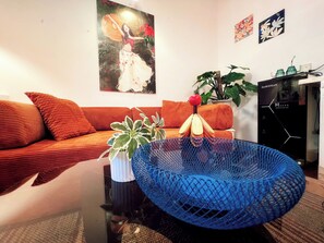 Een woonkamer met een bank, een blauwe geweven stoel, een potplant en een schilderij aan de muur.