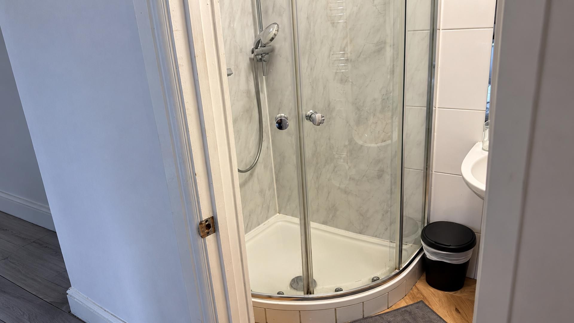 A glass-enclosed shower with a white tiled wall and a small trash bin beside it.