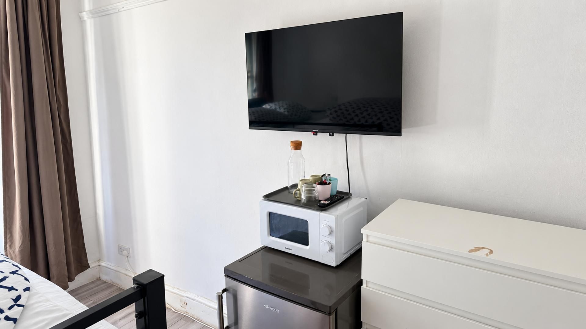 A hotel room with a bed, a white dresser, a microwave, a small fridge, and a flat-screen TV mounted on the wall.