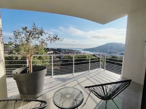 A balcony with a potted tree, a glass table, and two chairs, overlooking a coastal town and a mountain. - Luxury villa with ocean and mountain views (Port-Vendres)