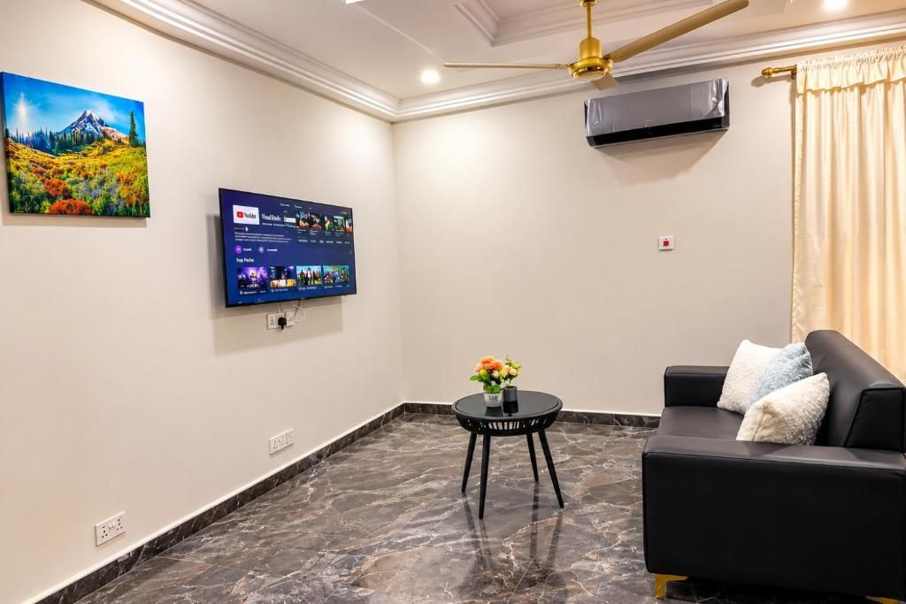 En moderne stue med et fladskærms-tv monteret på væggen, en sort lædersofa, et lille rundt bord med en vase blomster og en loftventilator.