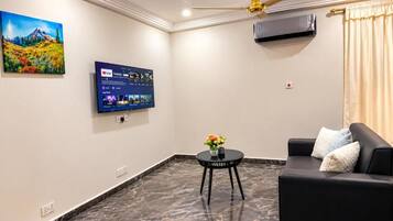 En moderne stue med et fladskærms-tv monteret på væggen, en sort lædersofa, et lille rundt bord med en vase blomster og en loftventilator.