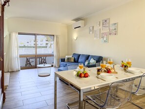 A dining area with a table set for two, a sofa in the background, and a balcony with a view of buildings. - #406 Modern 1-Bedroom Apartment with Pool | Near the Oura Strip (Albufeira)