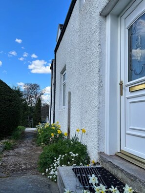 A white building with a door and a window, a stone pathway, and yellow flowers. - Charming cottage close to the Cairngorms (Ballindalloch)