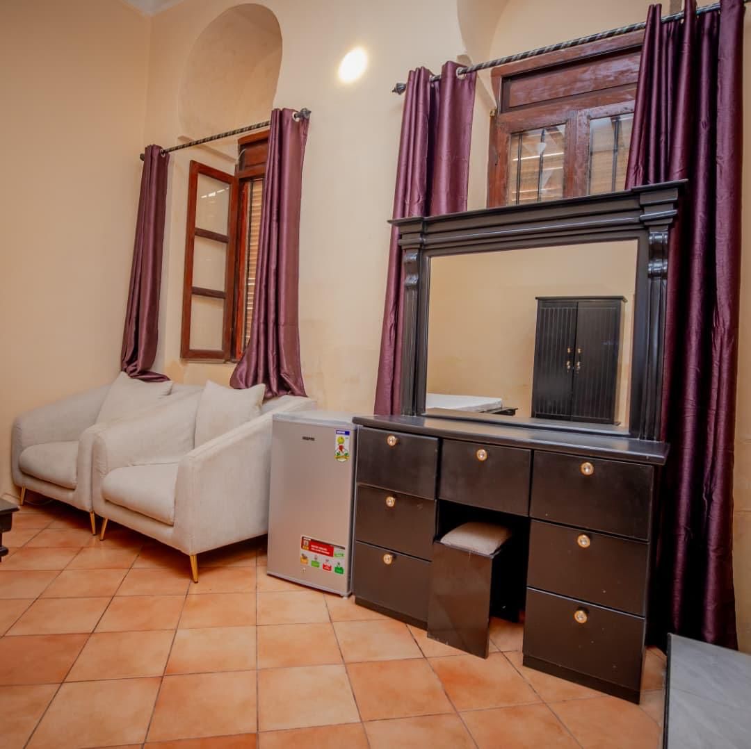Una habitación con sofá, refrigerador pequeño, cómoda con cajones y espejo sobre un mueble.