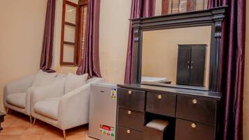 Una habitación con sofá, refrigerador pequeño, cómoda con cajones y espejo sobre un mueble.