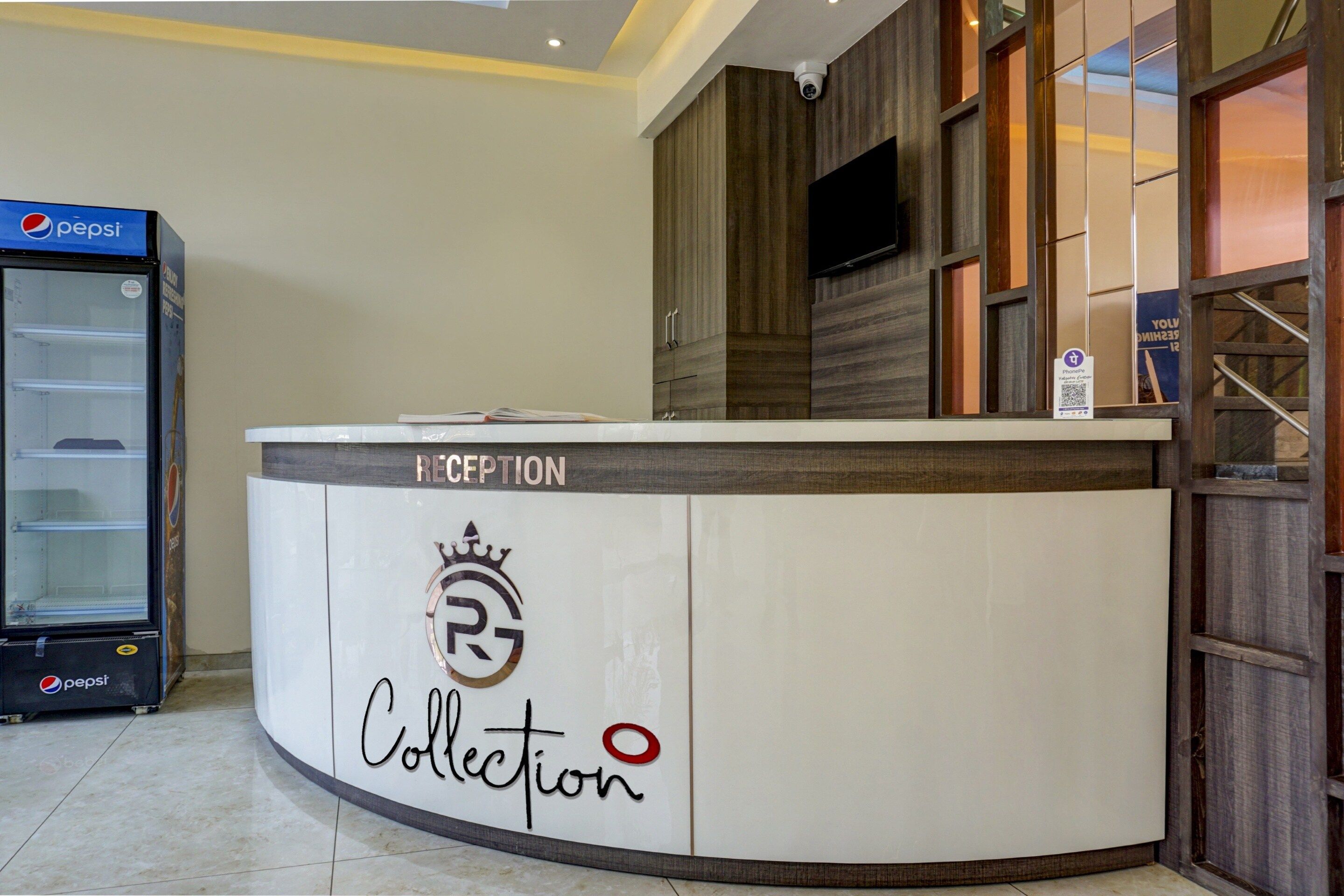 Un mostrador de recepción con un logotipo y la palabra 'Collection', un refrigerador Pepsi y una cámara de seguridad en la pared.
