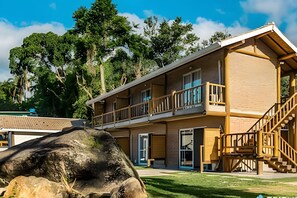 A two-story building with a balcony on the second floor, a staircase on the right, and a large rock in the foreground. - Recanto da Feiticeira (Ilhabela)