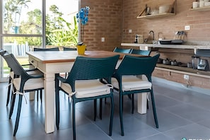 A modern kitchen with a brick wall, a dining table with chairs, and a view of greenery outside. - Recanto da Feiticeira (Ilhabela)