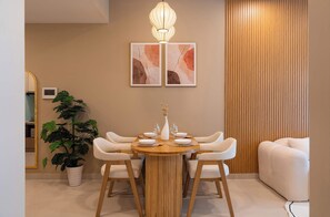 A dining area with a round wooden table, white upholstered chairs, a potted plant, and two abstract paintings on the wall. - High-Floor 2BR + 1BA | District 1 | Central City (Thành phố Hồ Chí Minh)