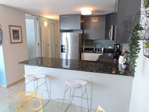 A modern kitchen with a stainless steel refrigerator, a built-in oven, and a coffee machine. There are two bar stools and a small dining table. - Seaside 3E - Luxury 2Br Apt on the Beach - Ocean View - Steps to Paradise (Carolina)
