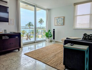 A living room with a sofa, a coffee table, a TV, and a balcony with a view of palm trees and the sea. - Seaside 3E - Luxury 2Br Apt on the Beach - Ocean View - Steps to Paradise (Carolina)