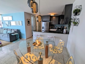 A modern living room with a glass dining table, a sofa, and a kitchen in the background. - Seaside 3E - Luxury 2Br Apt on the Beach - Ocean View - Steps to Paradise (Carolina)