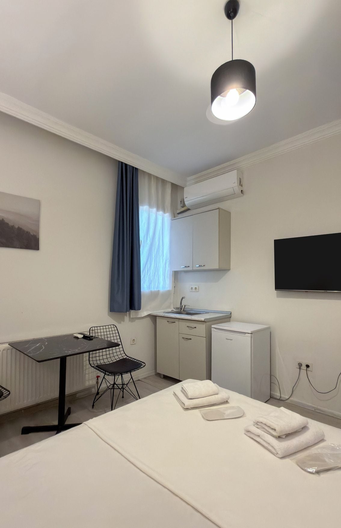 Habitación de hotel con cama, escritorio, silla, kitchenette pequeña y televisión.