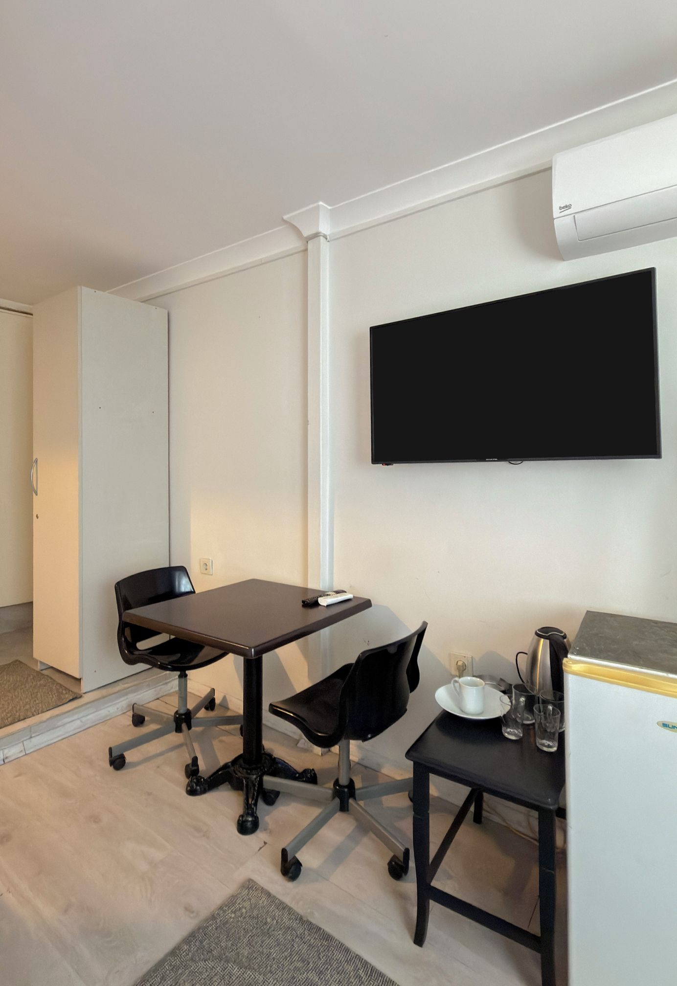 A modern room with a flat-screen TV mounted on the wall, a desk with a chair, and a small table with a coffee maker and cups.
