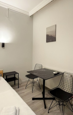 Une petite pièce, peu meublée, avec un lit, une table, deux chaises et un seul luminaire fixé au mur.