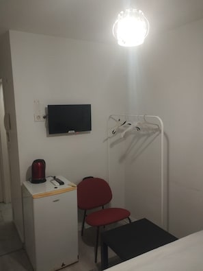 Une petite pièce simple, avec un téléviseur à écran plat fixé au mur, un bureau blanc avec une bouilloire et une télécommande, une chaise rouge, un banc et un étendoir à linge.
