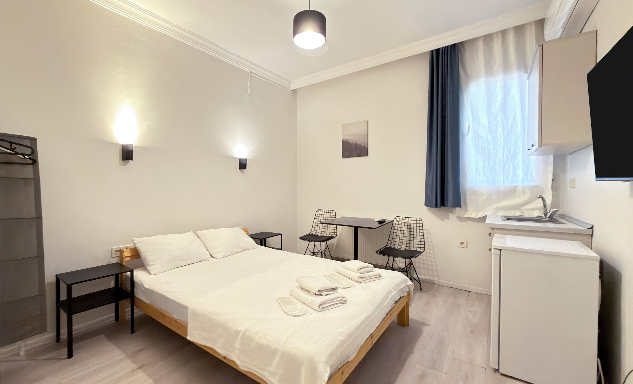 Una habitación de hotel con cama, mesitas de noche, escritorio con sillas, kitchenette pequeño y ventana con cortinas.