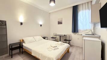 Una habitación de hotel con cama, mesitas de noche, escritorio con sillas, kitchenette pequeño y ventana con cortinas.