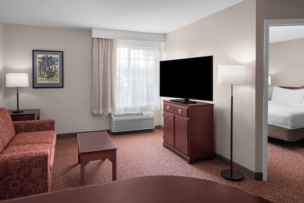 Ein Hotelzimmer mit Sofa, kleinem Tisch, Fernseher auf einem Schrank, Fenster mit Vorhängen und einem Bett.