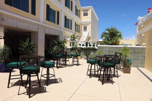 Eine Terrasse im Freien mit mehreren Tischen und Stühlen, umgeben von Gebäuden mit Balkonen und Topfpflanzen.