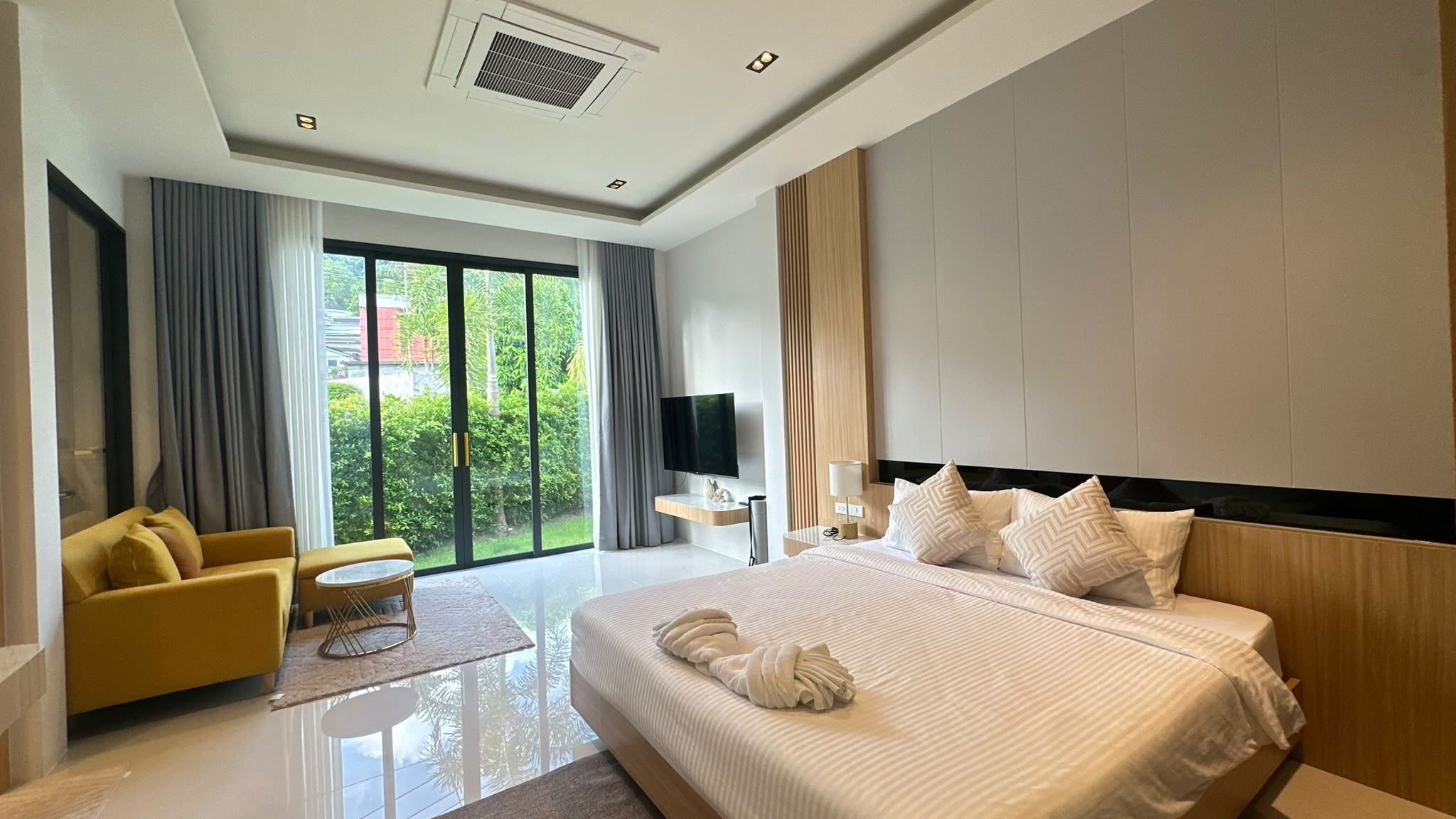 Een moderne hotelkamer met een groot bed, een zithoek met een gele bank, een flatscreen televisie en uitzicht op groen buiten.