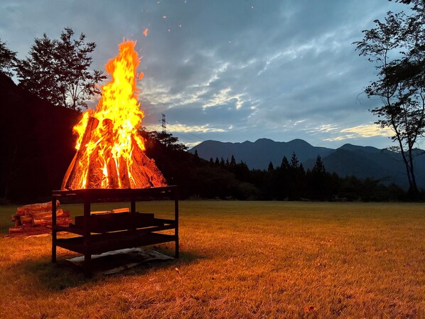 A campfire with a metal table in a grassy field with mountains in the background. - YOKOTE FOREST BASE (Aoki)