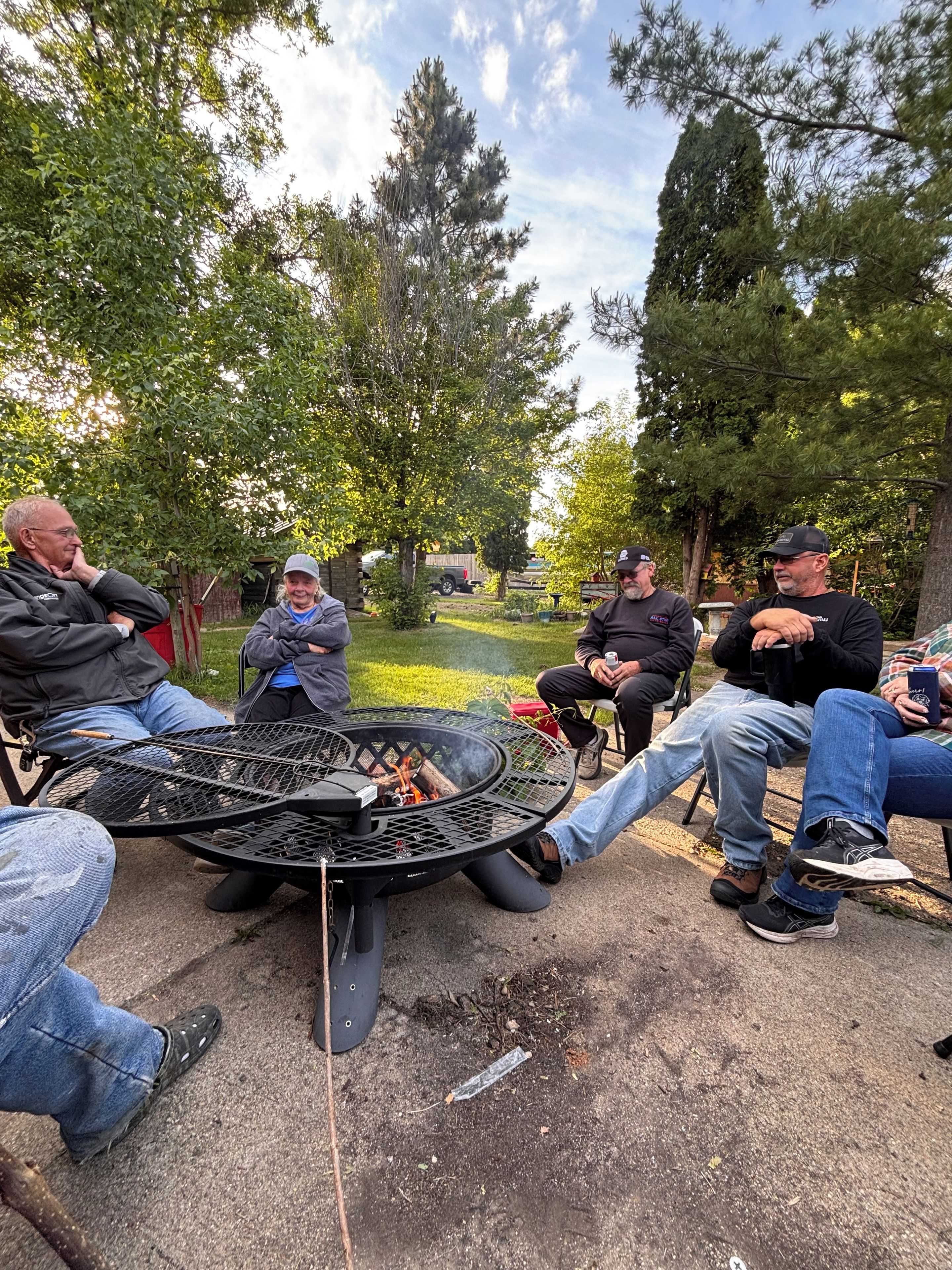 Eine Gruppe von Menschen sitzt um eine runde Feuerstelle im Freien mit einem Metallgrill, umgeben von Bäumen und Grünpflanzen.